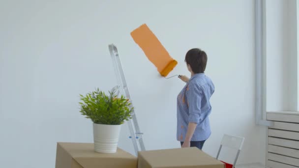 Düz yenileme konsepti. Orta yaşlı mutlu bir kadın boya silindirli, turuncu boyalı beyaz duvar boyuyor. - Video, Çekim