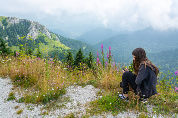 Νεαρή έφηβη κοπέλα που κοιτάζει αυτοτηλέφωνο στην κορυφή του βουνού ανάμεσα σε ροζ λουλούδια βουνού ενάντια στο πράσινο και θολό τοπίο του ουρανού - Φωτογραφία, εικόνα