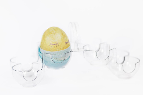 Velikonoční barevné kuřecí vejce v ochranné lékařské masce v chladničce na bílém pozadí. Koncepce prevence infekce kovid-19 koronavirem, během Velikonoc u pandemie - Fotografie, Obrázek