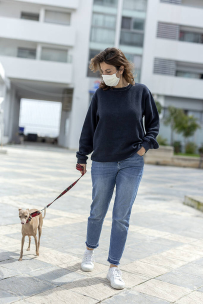 Woman with a protective mask against the coronavirus during the quarantine walking the dog inside an urbanization. Covid-19 concept - Фото, изображение
