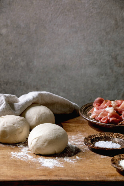 Dough for italian pizza napolitana cooking. Three balls of fresh homemade wheat dough, prosciutto and ingredients in ceramic plates above on wooden kitchen table. Home baking. - Fotó, kép