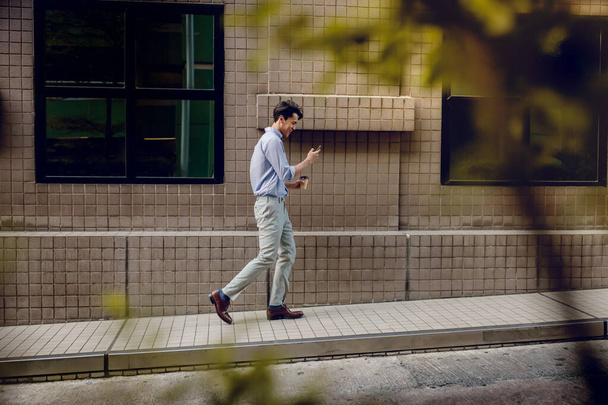 都会のビルを歩いている間に携帯電話を使用したり読んだりするカジュアルウェアの幸せな若いビジネスマン。現代人の生活様式。サイドビュー。全長 - 写真・画像