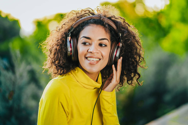 Zbliżenie afrykańskiej amerykanki słuchającej muzyki ze słuchawkami na zewnątrz. Młoda dziewczyna z kręconą fryzurą w żółtym cieszy się muzyką w parku. - Zdjęcie, obraz