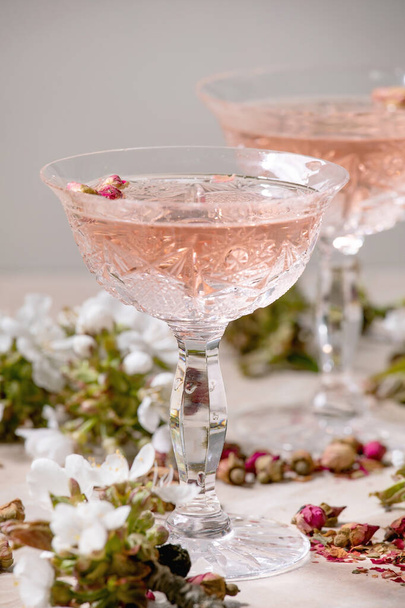 Křišťálové sklenice růžového šampaňského, moštu nebo limonády se suchými růžovými poupaty. Třešňové větve nad námi. Růžové mramorové pozadí - Fotografie, Obrázek