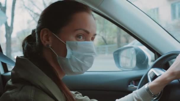 Beautiful Caucasian girl in blue protective medical face mask hold a black steering wheel, the driver looks around, checks for the absence of other cars. Brunette woman turns her head left and right. - Footage, Video