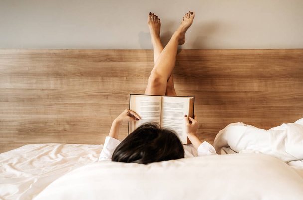 Ázsiai nő fekszik, keresztbe tett lábakkal, könyvet olvasott az ágyon. - Fotó, kép