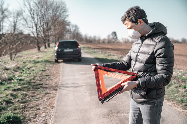 joven instala un triángulo de advertencia naranja en la carretera delante de un coche roto. Protección contra el coronavirus mediante respirador
 - Foto, imagen