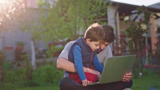 Een goede dag voor een vader en zijn schattige mooie jongen ze zitten op het gras in de tuin en chatten samen over de toekomst ze met behulp van de laptop om wat notities te maken over het kind dromen - Video