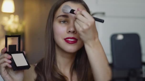 Portrait shot of the young pretty female beauty video blogger teaching how to do makeup and doing her forehead and cheeks with powder in front of the camera. - Materiaali, video