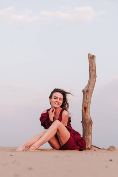 Girl sitting in the desert in the wind in a red dress near an old tree - Photo, Image
