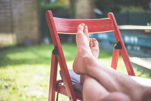 Young man is relaxing at home. Resting feet on a red chair, own garden in the blurry background - 写真・画像