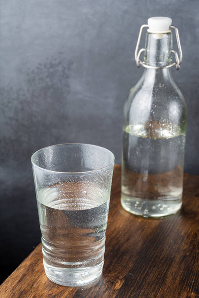 Nahaufnahme von feuchter Glasflasche und Glasflasche mit Wasser, auf dunklem Holztisch und grauem Hintergrund, vertikal, mit Kopierraum - Foto, Bild