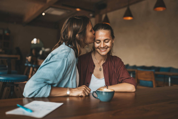 Happy young woman kissing her girlfriend, sitting in a cafe. Happy lesbian couple in love. - 写真・画像