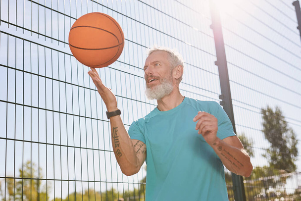 Jouer au basket à l'extérieur. Heureux homme âgé en vêtements de sport tournant une balle de basket-ball sur son doigt tout en se tenant debout sur le terrain de basket-ball extérieur
 - Photo, image