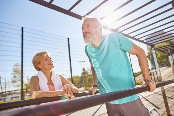 Senior family working out at outdoor gym. Mature bearded man in sportswear doing push ups on parallel bars. Active senior couple exercising in the morning - Fotó, kép