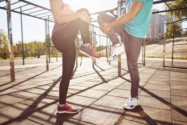 Stretching exercises. Cropped photo of man and woman in sportswear warming up together while standing at the stadium. They are stretching legs. Active couple doing sport outdoors - Фото, зображення