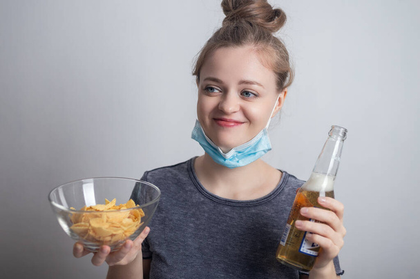 Hymyilevä nuori valkoihoinen nainen tyttö naamiossa juo pullon olutta ja pitää perunalastuja
 - Valokuva, kuva