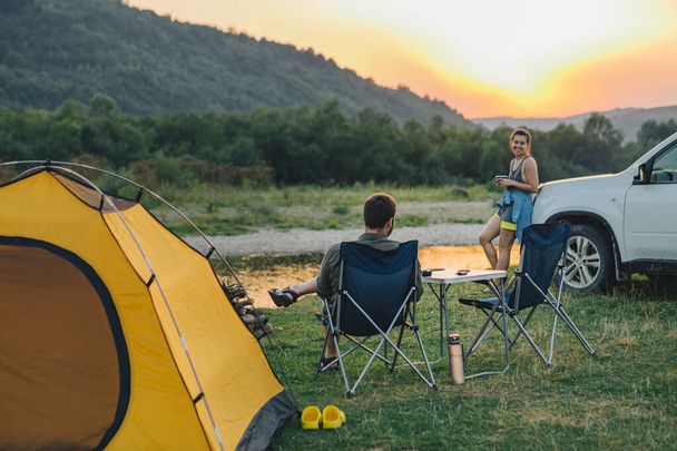 夫婦は山の中の川の上の夕日を見てキャンプチェアに座っていました。ハイキングのコンセプト. - 写真・画像