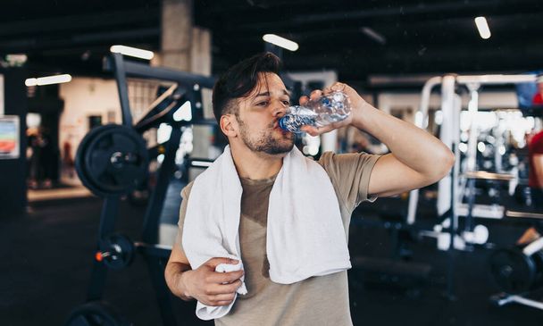 Young exhausted man in fitness gym after successful workout wipes sweat from the forehead with white towel while holding water bottle in other arm. - Photo, Image
