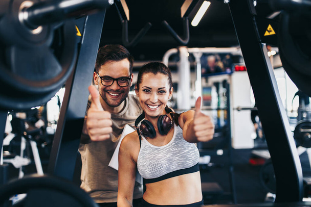 Young fit and attractive woman working out in modern gym together with her personal fitness instructor or coach. They looking at camera and smiling while showing thumb up. - Fotó, kép