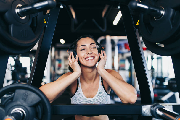 Young attractive woman after successful workout in modern fitness gym dancing, smiling and singing. She listening to music with bluetooth headset and smart phone. - Fotografie, Obrázek