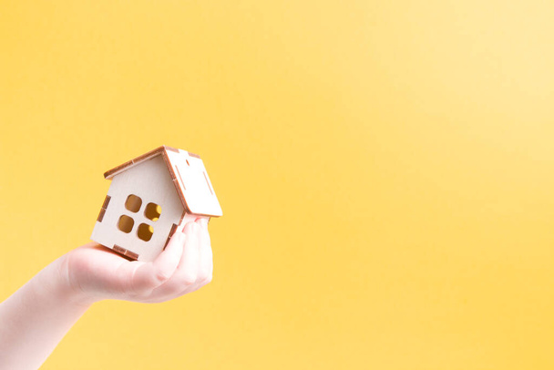 children's hand holds a small model of a wooden house, yellow background, copy space - Fotó, kép