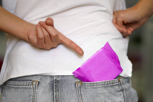 Женщина указывает пальцем на менструальную подушку в кармане джинсов. Женская гигиеническая прокладка в джинсовом кармане
. - Фото, изображение
