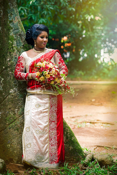 Kandy, Sri Lanka. 24 lipca 2016: Indyjska panna młoda w parku niedaleko drzewa w Kandy na Sri Lance. Nosi typową indyjską suknię ślubną. Z bukietem kwiatów w rękach. - Zdjęcie, obraz
