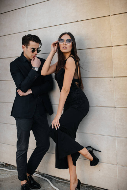 A beautiful, stylish pair of young people in black clothes and glasses stand against the background of an office building in the sunset. Fashion and style - Foto, immagini