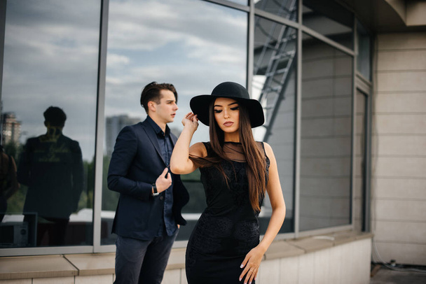 A beautiful, stylish pair of young people in black clothes and glasses stand against the background of an office building in the sunset. Fashion and style - Fotoğraf, Görsel