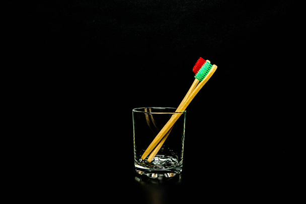 Dental and oral care products. Hygiene of teeth and oral cavity Two wooden toothbrushes in a glass cup on a black background. - Фото, зображення