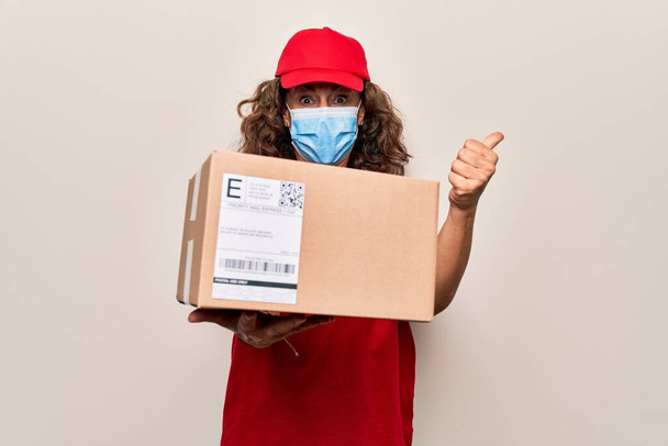 Middle age delivery woman wearing coronavirus protection mask and holding cardboard package pointing thumb up to the side smiling happy with open mouth - Photo, Image