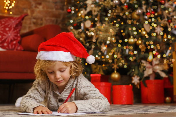 Little curly boy writes letter to Santa near christmas tree - Фото, зображення