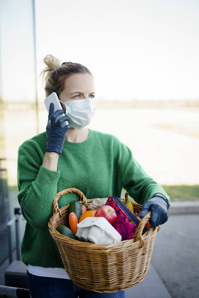 Pretty woman with face mask and green pullover and black gloves makes a phone call after shopping and is holding her shopping basket - Fotoğraf, Görsel