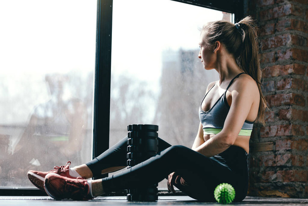 A girl in sportswear is sitting in the studio on the floor after a tense workout, next to her are a massage cylinder and myofascial ball. - Photo, image