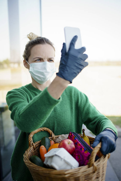 Pretty woman with face mask and green pullover and black gloves makes a phone call after shopping and is holding her shopping basket - Φωτογραφία, εικόνα