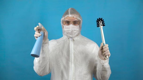 A young man in a protective suit with a toilet brush and a dispenser in his hands. Protection against virus outbreaks. In an antibacterial suit on a blue background. - Fotoğraf, Görsel