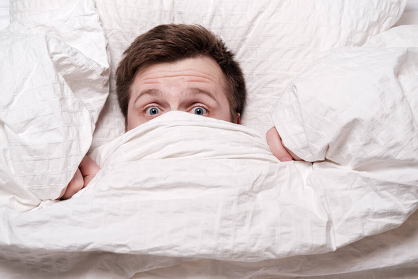 De man werd wakker uit een nachtmerrie, bedekte mond en neus met een deken en kijkt bang. Concept van slapeloosheid. - Foto, afbeelding