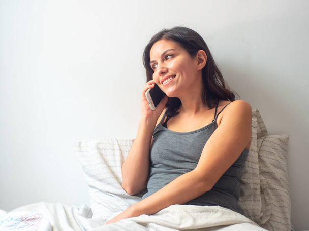 Jeune femme hispanique aux cheveux bruns reste à la maison et parle souriant au téléphone dans son lit en quarantaine
 - Photo, image