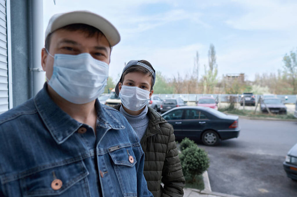 άνδρας και έφηβος αγόρι ποζάρουν κοντά σε τοίχο και κλειστή πόρτα πολυόροφων κτιρίων με διαμερίσματα, μια κατοικημένη περιοχή, μια ιατρική μάσκα στα πρόσωπά τους προστατεύει από ιούς και σκόνη - Φωτογραφία, εικόνα