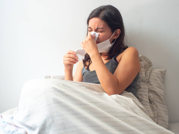 Νεαρή καστανή ισπανόφωνη γυναίκα είναι άρρωστη, φυσάει τη μύτη της και ξεκουράζεται στο κρεβάτι σε καραντίνα στην Καραντίνα. - Φωτογραφία, εικόνα