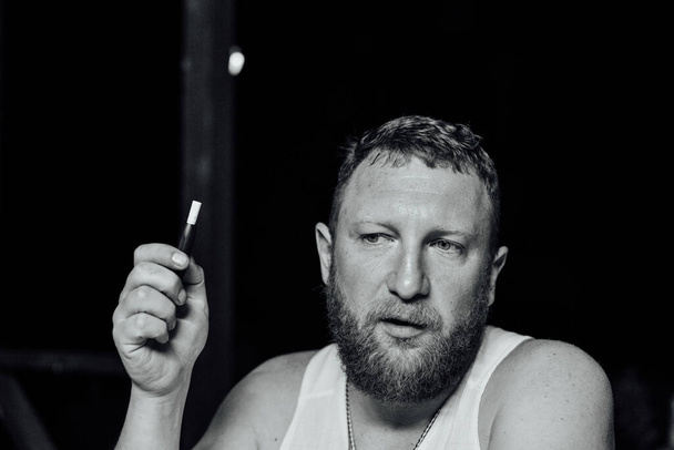 portrait of young grumpy bearded red hair man with sigaret horizontal. A man with a red beard smokes. The guy smokes an electronic cigarette. A man in a white T-shirt. Relaxation. Evening photo - Fotó, kép