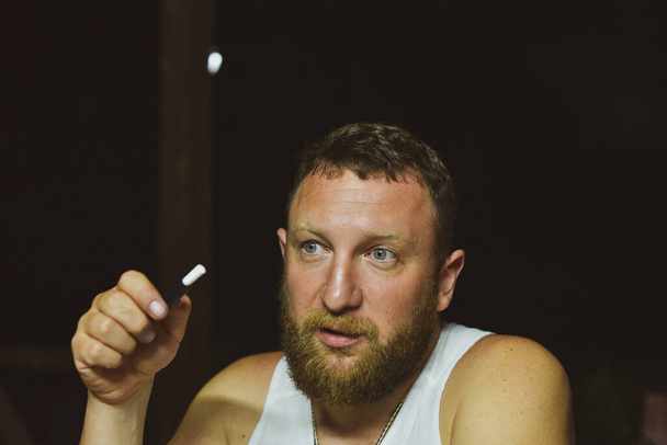 Huysuz, sakallı, sigara içen kızıl saçlı genç adamın portresi. Kızıl sakallı bir adam sigara içiyor. Adam elektronik bir sigara içiyor. Beyaz tişörtlü bir adam. Rahatlama. Akşam fotoğrafı. - Fotoğraf, Görsel