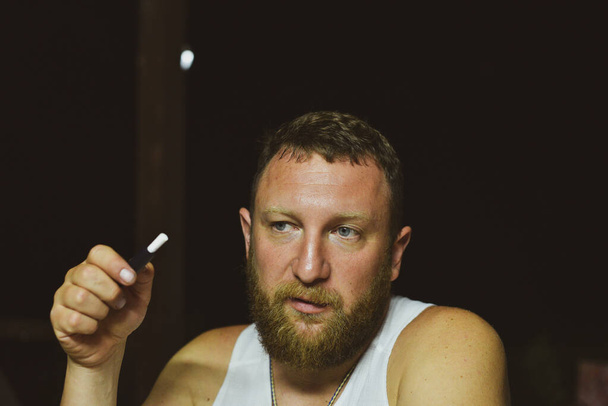 Porträt eines jungen mürrischen Mannes mit rotem Bart und horizontalem Sigaret. Ein Mann mit rotem Bart raucht. Der Mann raucht eine elektronische Zigarette. Ein Mann im weißen T-Shirt. Entspannung. Abendfoto - Foto, Bild