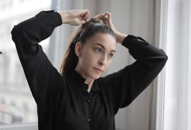 A beautiful closeup of a girl banding her ponytail in a black shirt against a blurry background - Foto, Imagen