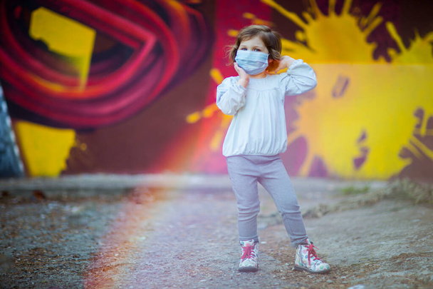 uma menina pequena, bonita, em uma máscara médica, fica na rua
 - Foto, Imagem