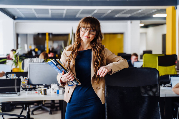 Smart girl with glasses, wearing classic office clothes, stands in coworking space, leaning her hand on a chair and looking to the camera. Other freelancers working on computers on  background - 写真・画像