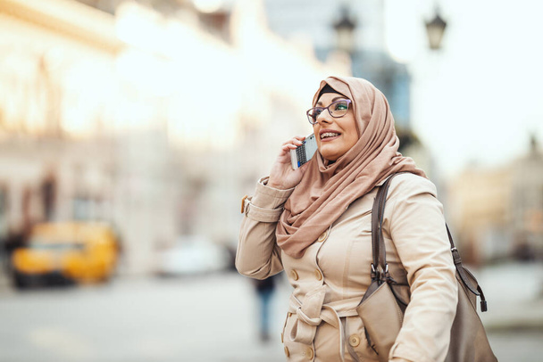 Mujer musulmana de mediana edad usando hijab con una cara feliz de pie en el entorno urbano, tomando en su teléfono inteligente
. - Foto, imagen