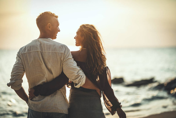 愛するカップルは、夕日の空の砂浜を楽しんで歩いています。彼らはお互いを見ている. - 写真・画像