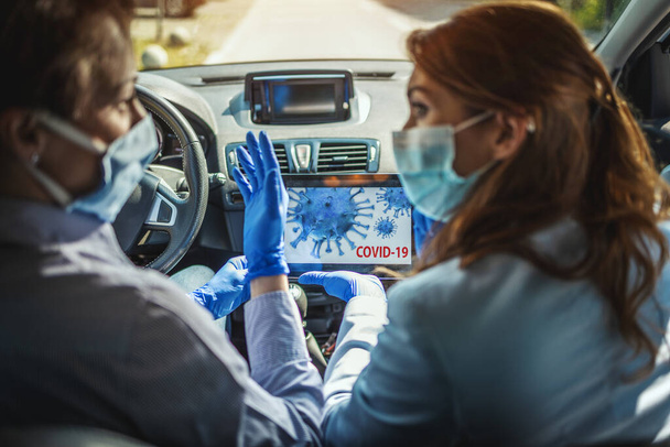 Young business women with protective masks on their faces are sitting in a car and talking about global crisis. They are showing each other an animation of a corona virus on a tablet. - Photo, Image